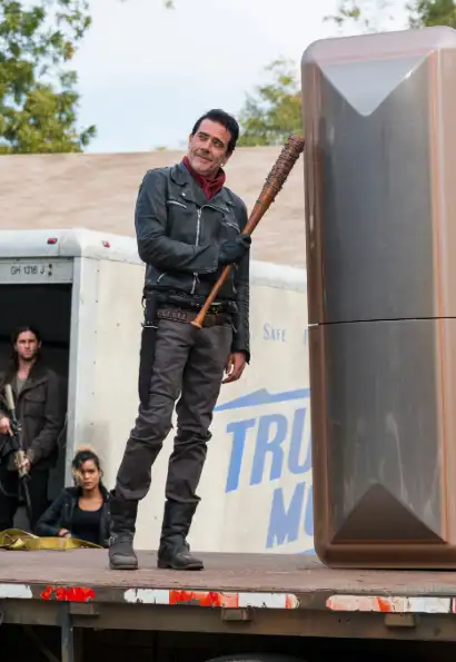 Jeffrey Dean Morgan als Negan in The Walking Dead. Er trägt eine schwarze lederjacke mit einem roten Halstuch, eine dunkle Jeans mit schwarzen Stiefeln. In seiner Hand hält er einen Baseballschläger, mit dem er auf einen Kühlschrank zeigt. Er steht auf einer Bühne.