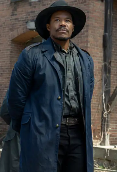 Gauis Charles als Perlie Armstrong in Dead City. Er trägt einen Hut und einen blauen Mantel 