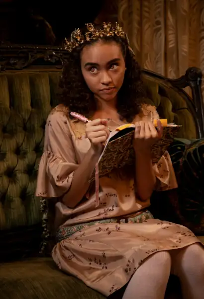 Brass Bailey als Claudia in Interview with the Vampire. Sie trägt eine Krone auf dem Kopf und schreibt in ein Buch. Sie trägt ein Kleid.