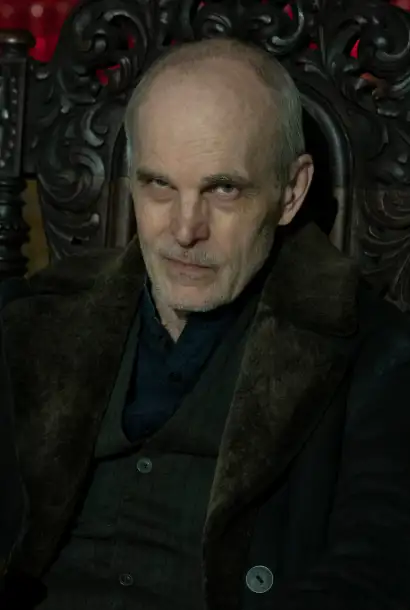Željko Ivanek als The Croat in Dead City. Er sitzt auf einem Thron und trägt einen Pelzmantel. 