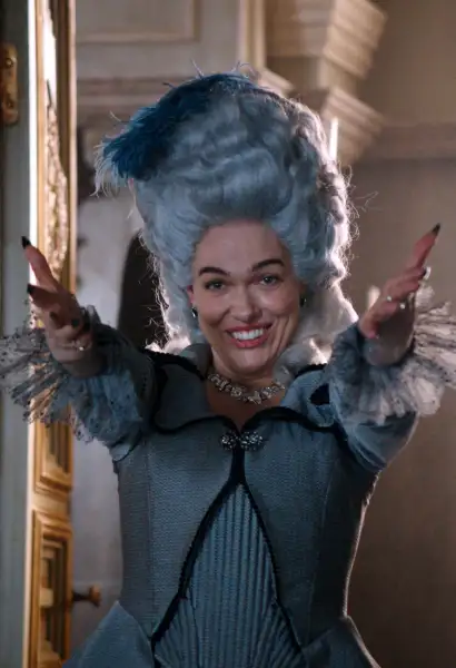 Verena Altenberger als Marie Antoinette in Mozart/Mozart. Sie trägt eine weiße Perücke, ein weißes dekandentes Kleid. Ihre Hände sind willkommend ausgestreckt und sie trägt ein lächeln. Ihre Nägel sind lackiert und sie trägt eine teure Kette.