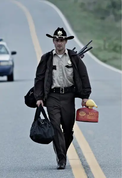 Andrew Lincoln als Rick Grimes in The Walking Dead. Er trägt eine Polizeiuniform, auf seinem Rücken ist ein Beutel voller Schusswaffen. In seiner linken Hand ist eine Tragetasche, in seiner rechten trägt er einen Werkzeugkoffer.