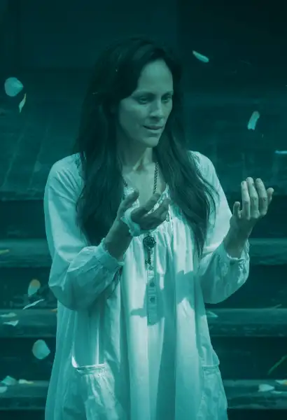 Anabeth Gish als Deirdre Mayfair inMayfair Witches. Sie starrt begeistert auf ihre Hände, um sie herum fliegen Schmetterlinge.Sie trägt ein weißes Nachthemd.
