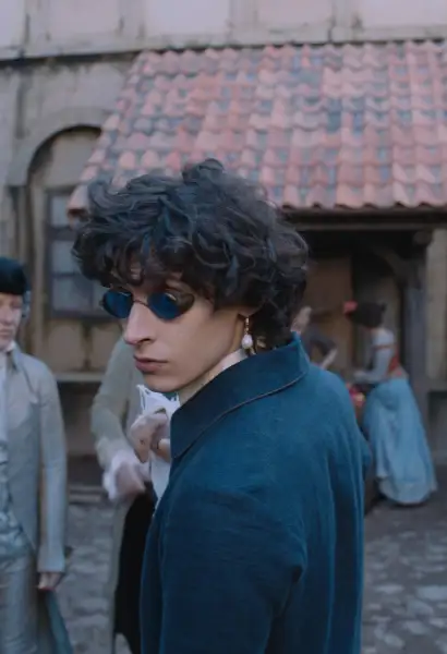 Eren M. Güvenci als Wolfgang Amadeus Mozart in Mozart/Mozart. Er trägt eine modische Brille, eine blaue Jacke aus dem 18. Jahrhundert.  Im Hintergrund ist ein Haus zu sehen.
