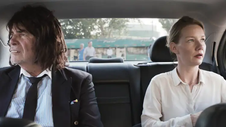 Peter Simonischek als Toni Erdmann und Sandra Hüller als Ines in Toni Erdmann. Sie sitzen in einem Taxi, er trägt einen Anzug und Perücke, sie trägt ein weißes Hemd.
