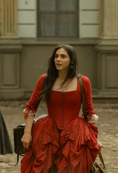 Havana Joy Braun als Marie Anne in Mozart/Mozart. Sie trägt ein rotes Kleid aus dem 18. Jahrhundert und trägt Koffer mit sich.