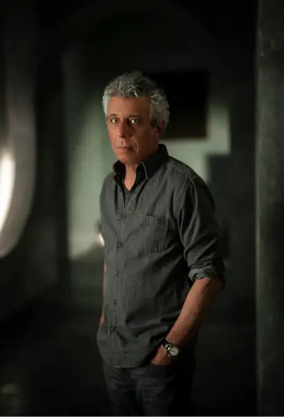 Eric Bogosian als Daniel Molloy in Interview with the Vampire. Er trägt ein graues Hemd und dunkle Jeans. Auf seinem Arm trägt er eine Uhr.