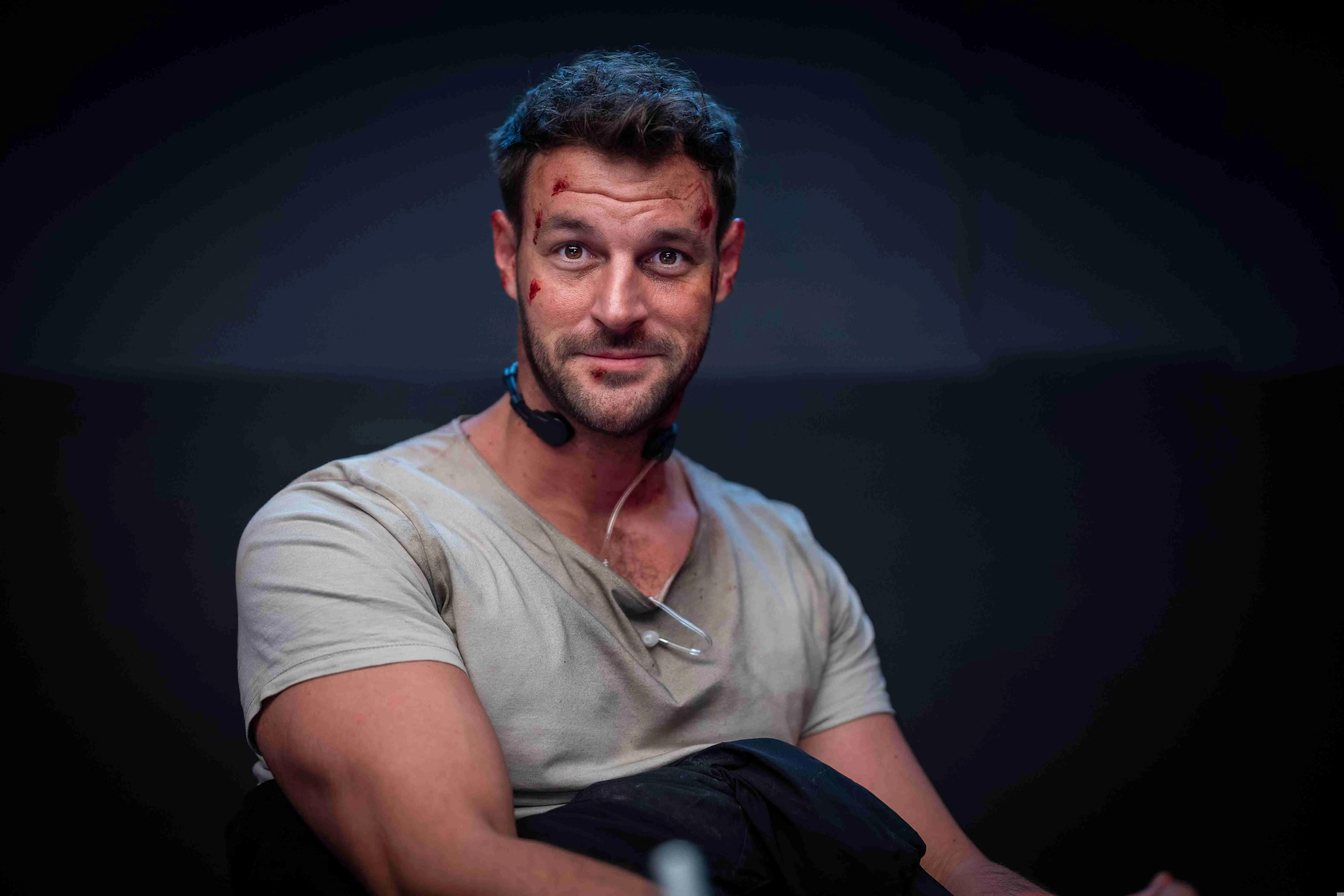 Paul Pizzera als Flo Kienzl in Neo Nuggets, sitzend in grauem T-Shirt, mit skeptischem Blick in einer dunklen Szene des österreichischen Action-Comedy-Films.