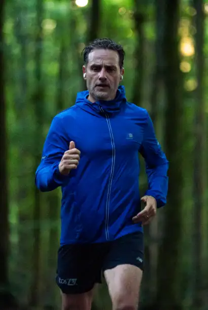 Andrew Lincoln als John in Coldwater in einem blauen Trainingsanzug. Er läuft. Im Hintergrund ist in Wald zu sehen.