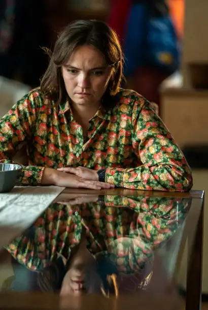 Abigail Lawrence als Moira-Jean in Coldwater. Sie trägt ein buntes Kleid. Ein Tisch im Bild speigelt sie wieder. Sie hat einen verstörten Blick.