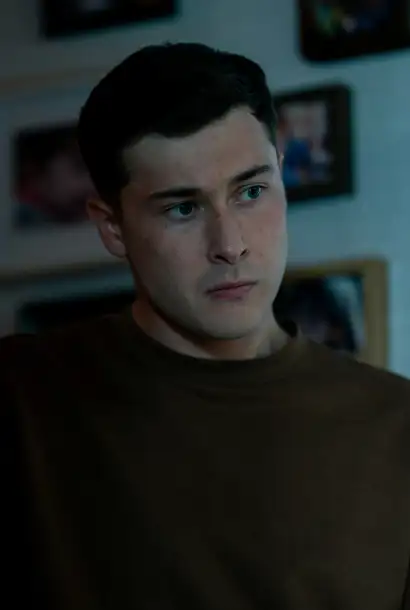 Samuel Bottomley als Cameron in Coldwater. Er trägt ein schwarzen Pullover. Sein Blick ist ernst. Im Hintergrund ist eine Wand zusehen an der Bilder hängen.