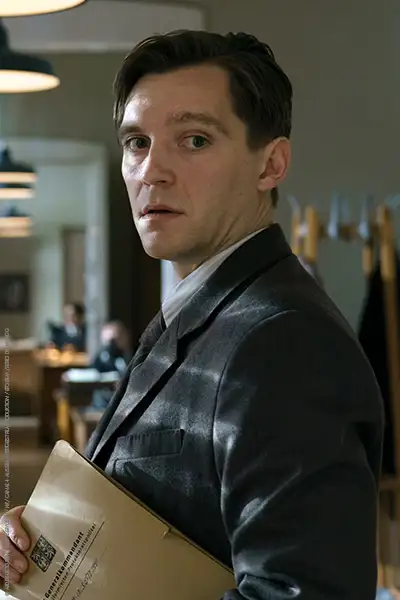 Jonas Nay als Jan Morava, einen Aktenordner unter den Arm geklemmt. Im Hintergrund ist ein Polizeibüro zu sehen.