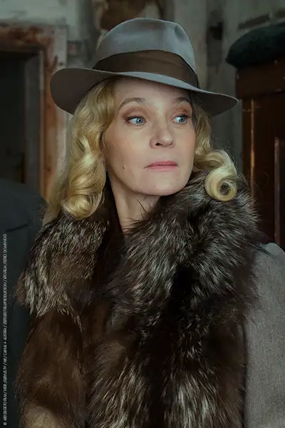 Jeanette Hain als Marleen Baumann in einem 40er Jahre Mantel mit Pelzbesatz, einem schief sitzenden Fedora-Hut und blonden Locken.