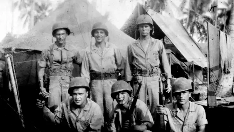Schwarz Weiß Foto von Soldaten in Unifrom im zweiten Weltkrieg. Im Hintergrund sind Zelte und Palmen zu sehen.
