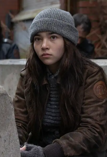 Mahina Napoleon s  in einem beanie und einer Jacke als Ginny in Dead City. Sie sitzt auf der Straße.
