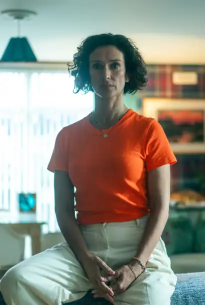 Indira Varma as Fiona in Coldwater in einem orangen T-Shirt und beiger Hose. Im Hintergrund erkennt man Inneneinrichtung eines Hauses.
