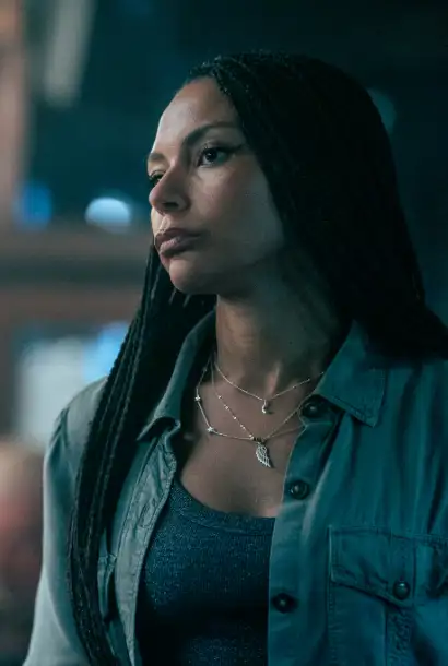 Lois Chimmiba als Catriona in Cold Water. Sie trägt eine Kette, ein grünes Hemd und ein dunkel blaues Shirt. Im Hintergrund ist verschwommen eine Bar zu sehen.