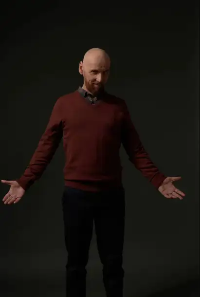 Ewen Bremner als Tommy in einem roten Pullover und schwarzer Hose. Er lächelt und hält die Arme weit Ausgestreckt. der Hintergund ist dunkel.