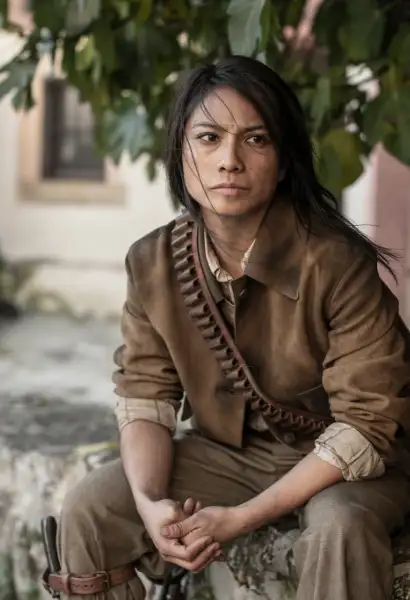 Alexandra Masangkay als Paz in The Walking Dead: Dary Dixon. Sie trägt eine Braune Jacke mit dunkelbraunen Hosen. Um Ihren Torso ist ein Munitionsgurt aus Leder umgehängt.