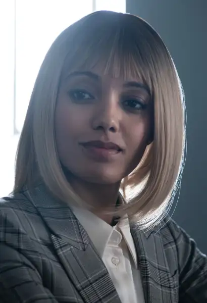 Maise Richardson-Sellers als Olive in Talamasca. Sie trägt eine Blonde Perrücke und einen grauen, gestreiften Anzug.