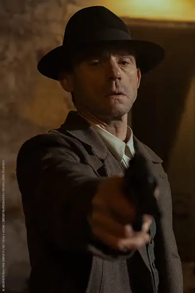 Gerhard Liebmann als Antonín Rypl im Mantel mit Hut, eine Pistole nach unten gerichtet.